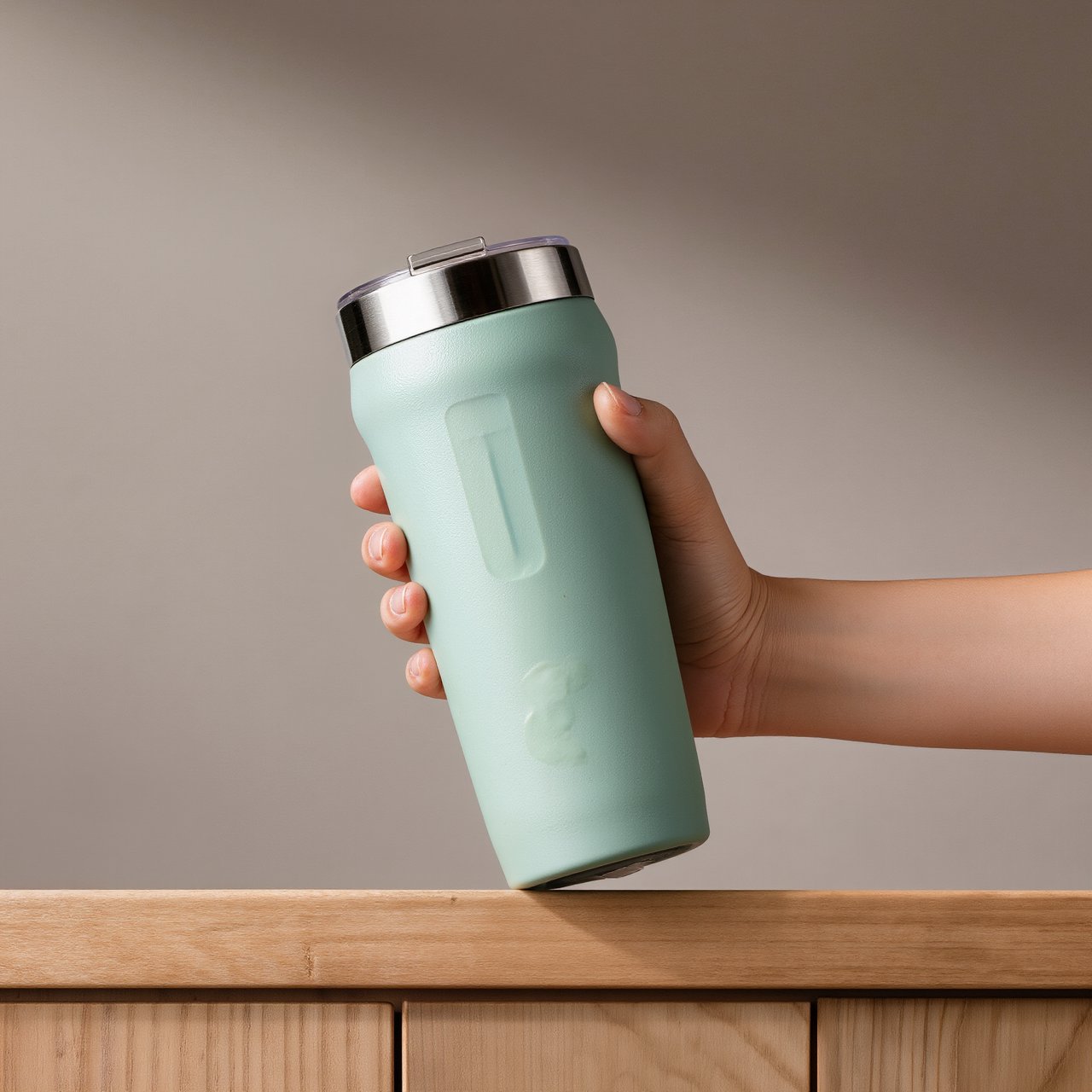An adult hand lifts an unbranded insulated travel mug, its matte sea-glass polymer body and brushed-steel rim catching softbox highlights above a pale oak counter; warm gray studio backdrop, broad diffused key and cool edge light, no face or lettering. — Kling v3