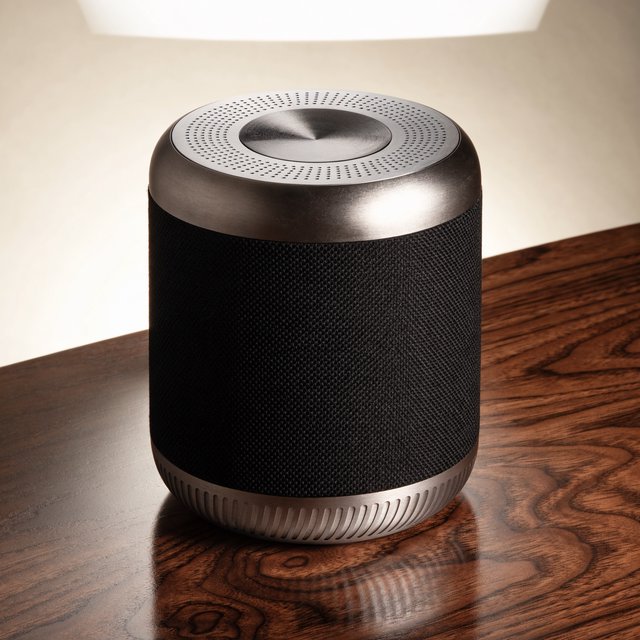 Commercial product photograph of an unbranded wireless speaker made from textured black fabric and satin aluminum, placed on a walnut tabletop against a warm cream background, illuminated by a large softbox and subtle backlight. — Recraft v4 Pro