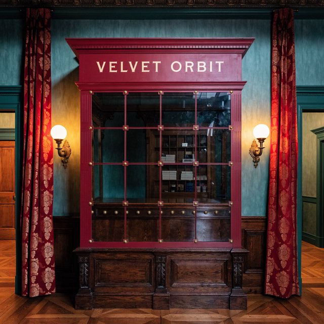 In a restored neighborhood cinema, brass sconces illuminate velvet curtains and a glass ticket booth; "VELVET ORBIT" is painted in ivory block letters across the booth’s burgundy fascia. — Recraft v4 Pro