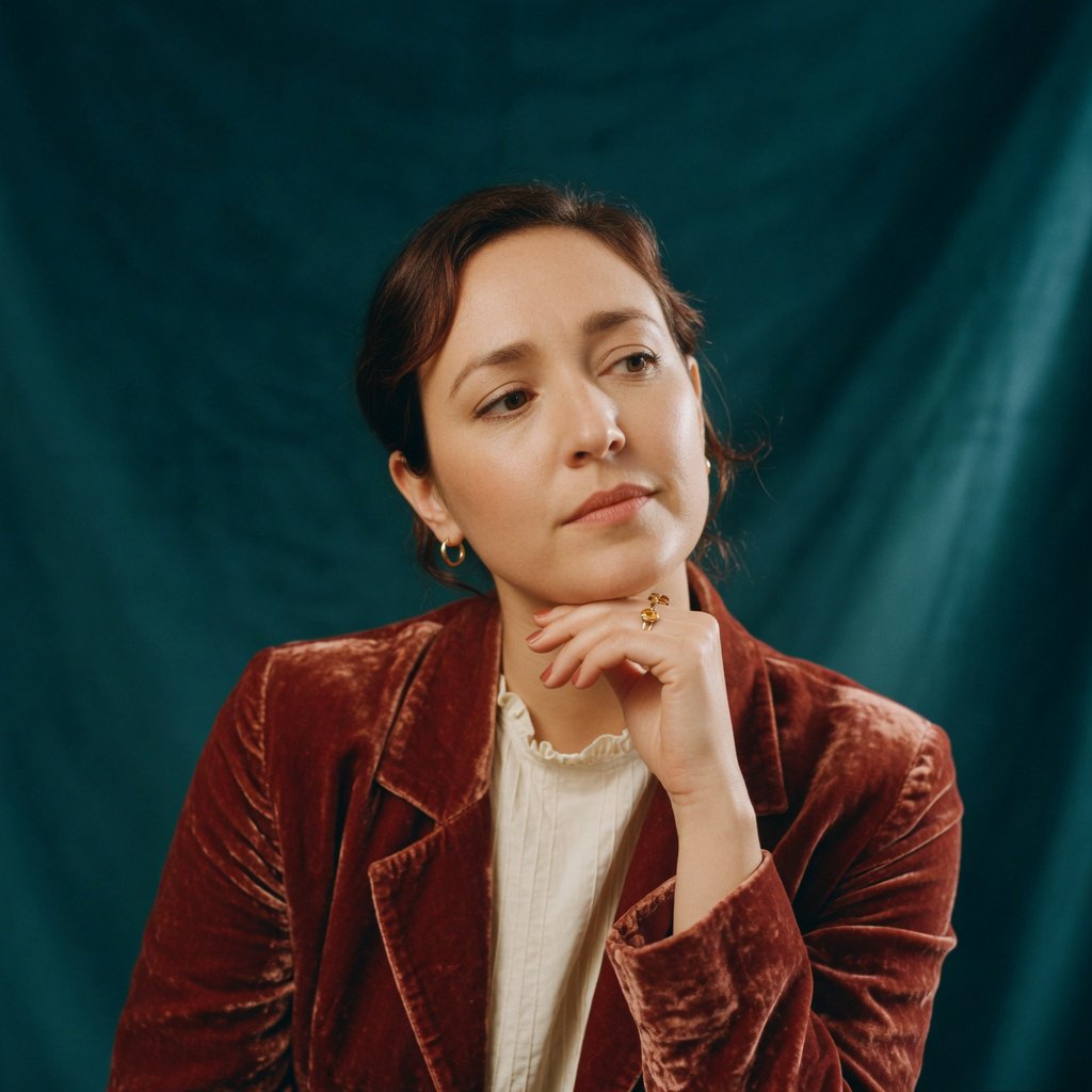 Portrait of an adult woman in her 30s wearing a rust-colored velvet jacket over an ivory blouse, one hand resting beneath her chin, thoughtful expression, deep teal studio backdrop, soft side lighting and gentle cheek highlights. — Grok Imagine Image Quality