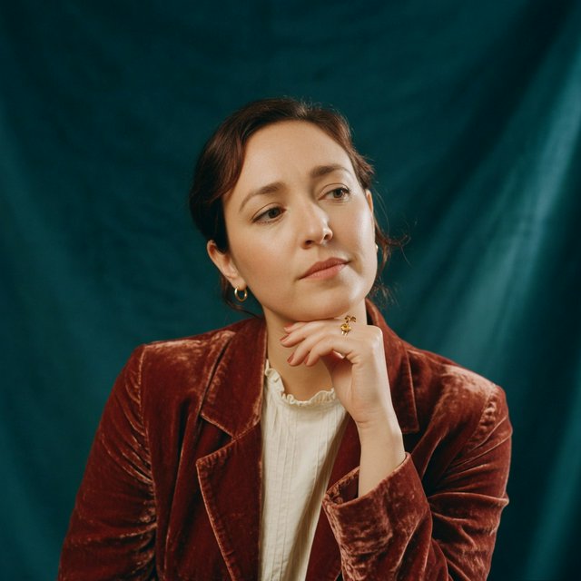 Portrait of an adult woman in her 30s wearing a rust-colored velvet jacket over an ivory blouse, one hand resting beneath her chin, thoughtful expression, deep teal studio backdrop, soft side lighting and gentle cheek highlights. — Grok Imagine Image Quality