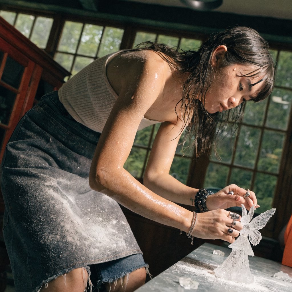 A pulchritudinous, gorgeous, exotic East Asian beauty, aged 24, inside an atelier, wearing a loose workshop apron, a tube top, and hot pants. Sweating profusely, she remains concentrated. She has a focused posture and expression as she looks at the crystalline object in the foreground that she is sculpting into a small, detailed fairy with high precision and clarity. Three-quarter-body mid-shot, Dutch angle. — Recraft v4