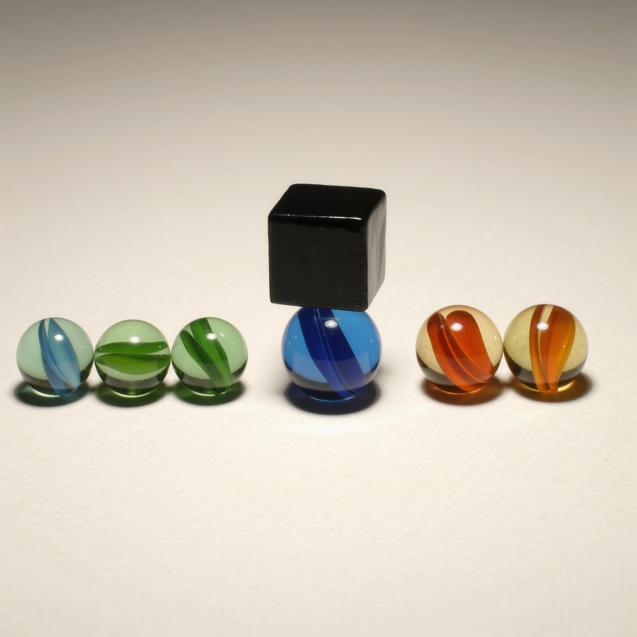 Exactly six glass marbles sit on a cream tabletop: three green marbles in a row on the left, two amber marbles in a row on the right, and one blue marble centered between the rows. A black cube sits above the blue marble, nothing else. — Kling v3 Omni