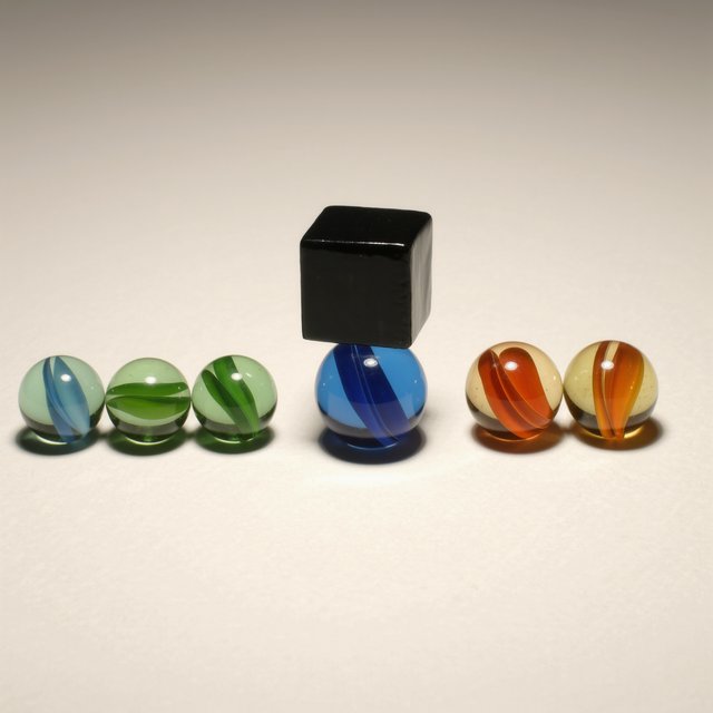 Exactly six glass marbles sit on a cream tabletop: three green marbles in a row on the left, two amber marbles in a row on the right, and one blue marble centered between the rows. A black cube sits above the blue marble, nothing else. — Kling v3 Omni