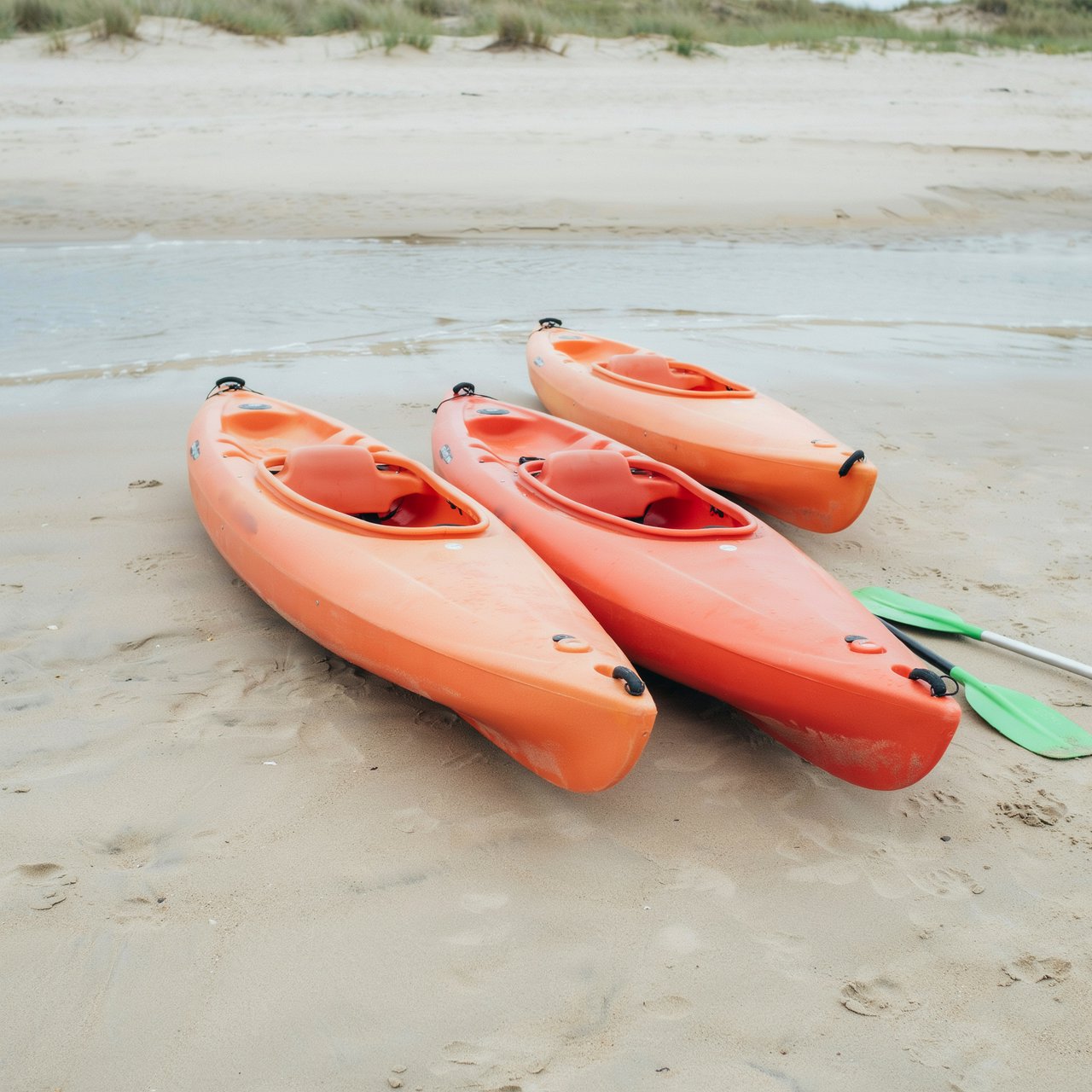 Exactly three orange kayaks rest on a sandy shore: two side by side in the foreground and one behind them near the water. A green paddle lies to the right of the kayaks, nothing else. — Ideogram v4