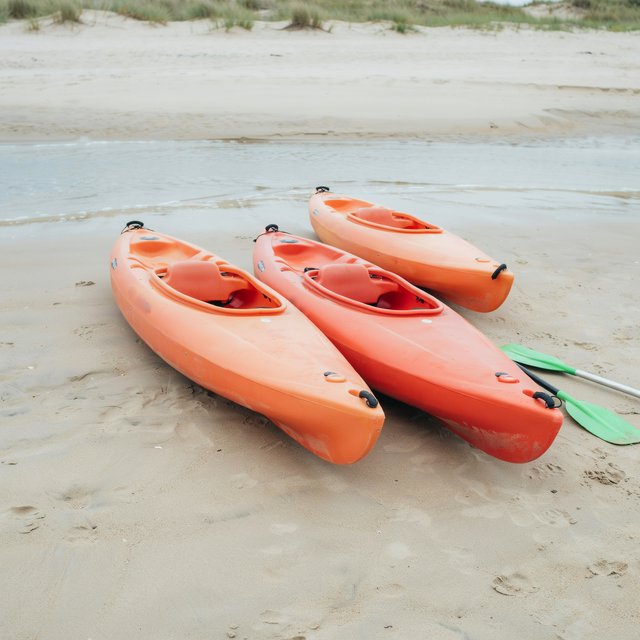 Exactly three orange kayaks rest on a sandy shore: two side by side in the foreground and one behind them near the water. A green paddle lies to the right of the kayaks, nothing else. — Ideogram v4