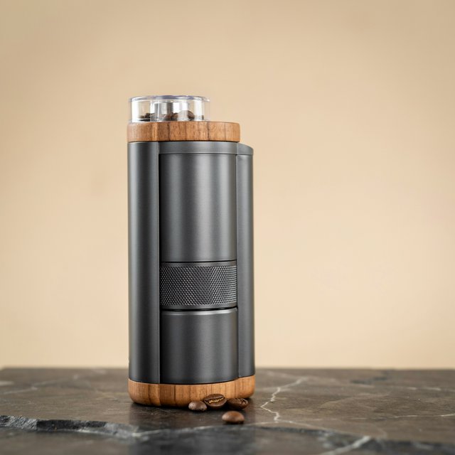 Commercial product photograph of an unbranded portable espresso grinder crafted from anodized aluminum with walnut accents, resting on a black stone surface against a warm beige background, illuminated by broad diffused studio light and a subtle edge highlight. — Ideogram v4
