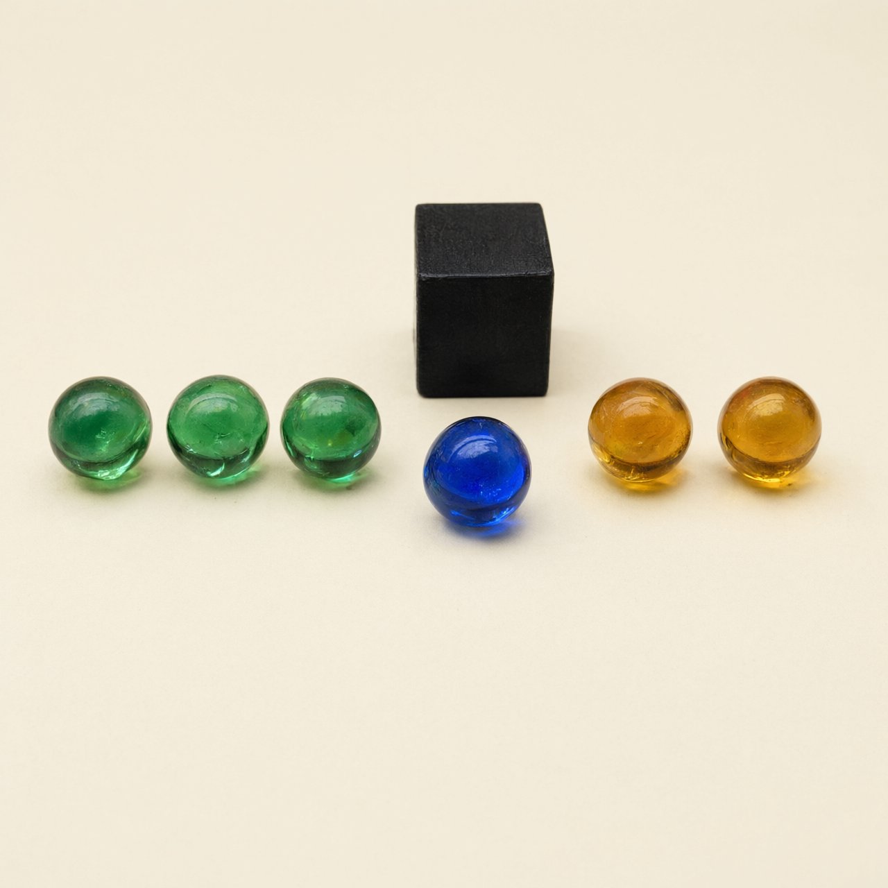 Exactly six glass marbles sit on a cream tabletop: three green marbles in a row on the left, two amber marbles in a row on the right, and one blue marble centered between the rows. A black cube sits above the blue marble, nothing else. — Qwen Image 3.0 Pro