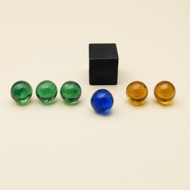 Exactly six glass marbles sit on a cream tabletop: three green marbles in a row on the left, two amber marbles in a row on the right, and one blue marble centered between the rows. A black cube sits above the blue marble, nothing else. — Qwen Image 3.0 Pro