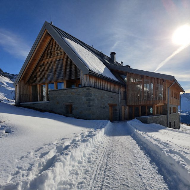 Exterior of a contemporary alpine research lodge in restrained regional modernism, rough granite walls, larch screens, steep standing-seam roof, crisp snow-bright morning light, viewed from below across a mountain footpath, sound structural geometry. — ImagineArt 1.5 Pro