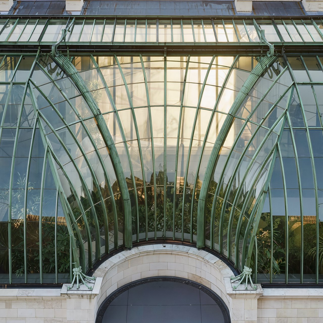 From a nearby rooftop, an Art Nouveau conservatory curves around a planted courtyard, its green copper ribs, clear glass, and pale stone base catching hazy morning light; orderly rooflines and believable supports show from above. — Kling v3