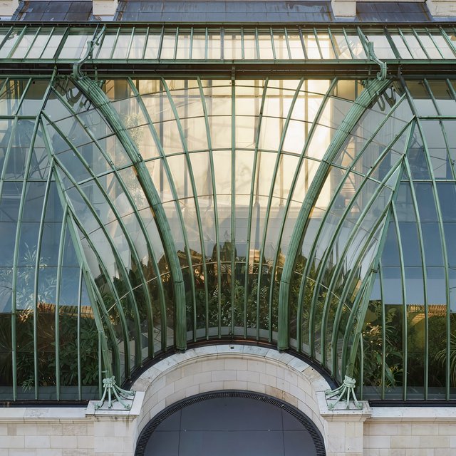 From a nearby rooftop, an Art Nouveau conservatory curves around a planted courtyard, its green copper ribs, clear glass, and pale stone base catching hazy morning light; orderly rooflines and believable supports show from above. — Kling v3