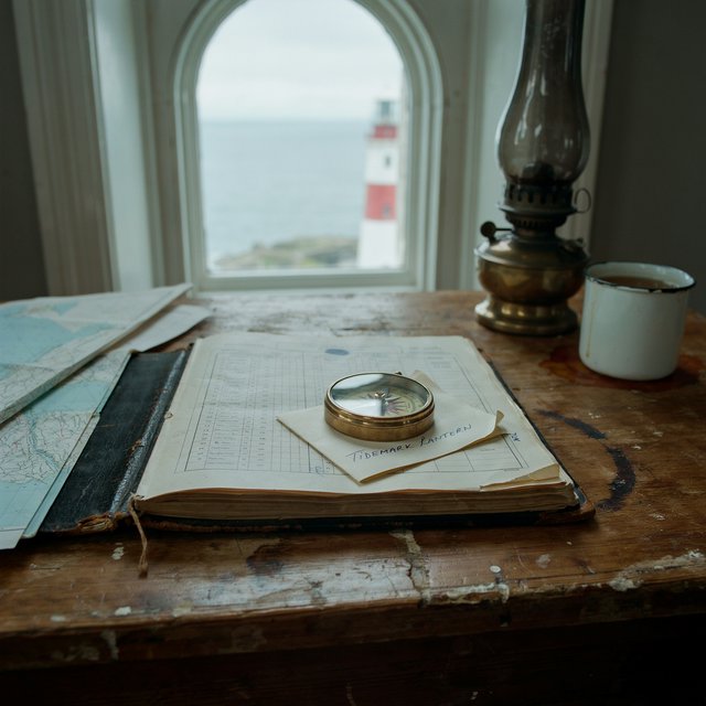Beside a cliffside lighthouse, a salt-speckled logbook lies open on a chart table; "TIDEMARK LANTERN" is handwritten in looping dark-blue ink across a cream note tucked beneath the compass, with no other legible writing. — Ideogram v4