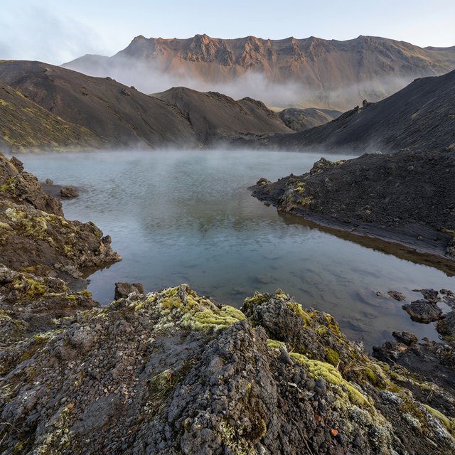 At first light, a rain-cleared volcanic caldera lake lies beneath thinning fog; pumice grains and damp lichen sharpen the near shore, while dark cinder slopes step back toward a distant rim. — Qwen Image 3.0 Pro