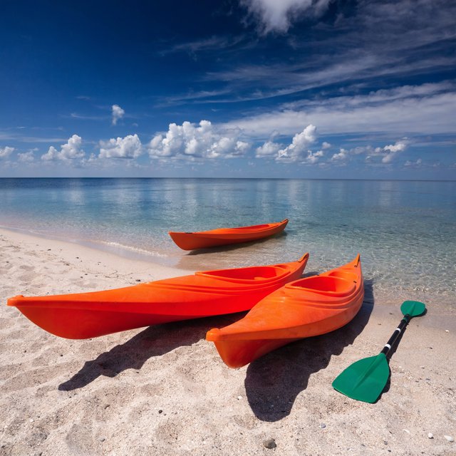 Exactly three orange kayaks rest on a sandy shore: two side by side in the foreground and one behind them near the water. A green paddle lies to the right of the kayaks, nothing else. — Recraft v4 Pro
