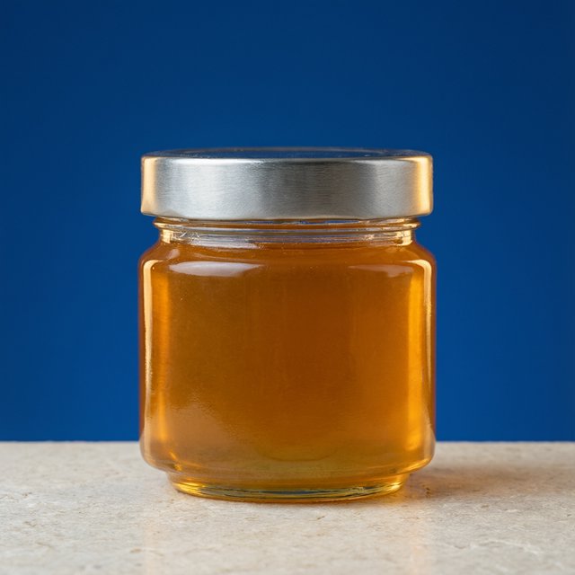 A squat unbranded honey jar with clear glass walls and a plain brushed-aluminum lid rests on honed pale limestone; broad diffused key light and warm rim light shape the commercial product image against a seamless cobalt-blue background, with no lettering. — Model 18