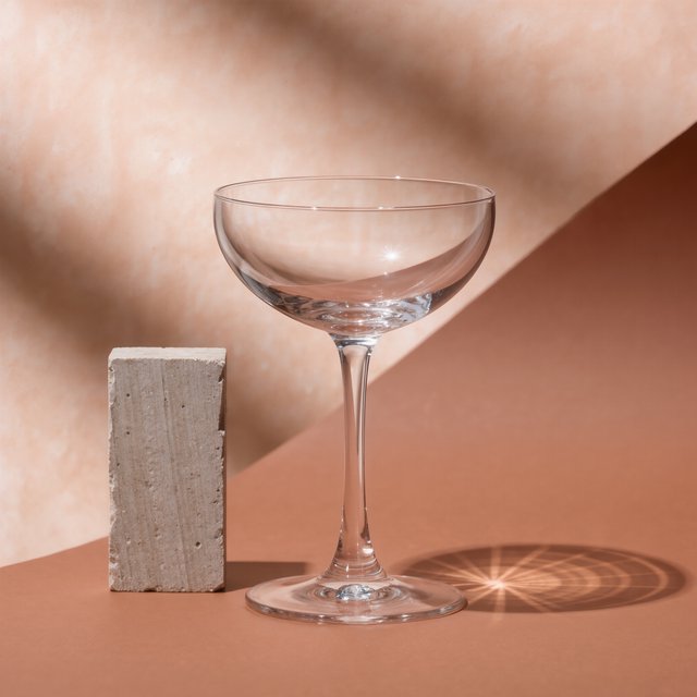 Beside a pale limestone block, an unbranded handblown cocktail coupe in clear soda-lime glass catches crisp window-shaped highlights; its curved bowl bends a muted apricot backdrop under broad diffused key light and narrow cool rim lighting, commercial product image, no lettering. — Kling v3 Omni