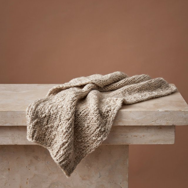 Soft folds of an unbranded chunky-knit wool throw rest on a pale travertine bench; its looped surface catches broad diffused key light and gentle side fill against a muted terracotta studio background, forming a clean commercial product image with no lettering. — Model 8
