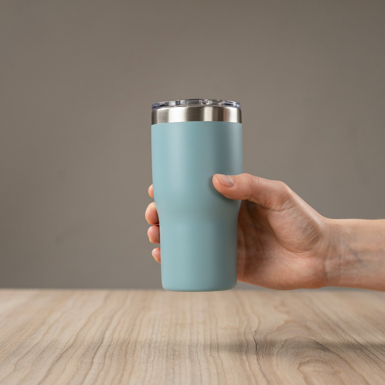 An adult hand lifts an unbranded insulated travel mug, its matte sea-glass polymer body and brushed-steel rim catching softbox highlights above a pale oak counter; warm gray studio backdrop, broad diffused key and cool edge light, no face or lettering. — Ideogram v4