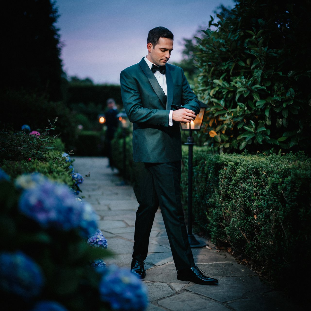 Along a garden path at violet dusk, an adult man in a midnight-green dinner suit adjusts one cuff beneath warm lantern glow, caught in a quiet pre-event pause with glossy leaves and elegant magazine polish. — Ideogram v4