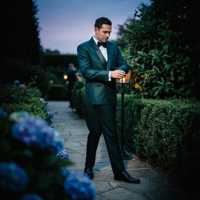 Along a garden path at violet dusk, an adult man in a midnight-green dinner suit adjusts one cuff beneath warm lantern glow, caught in a quiet pre-event pause with glossy leaves and elegant magazine polish. — Ideogram v4