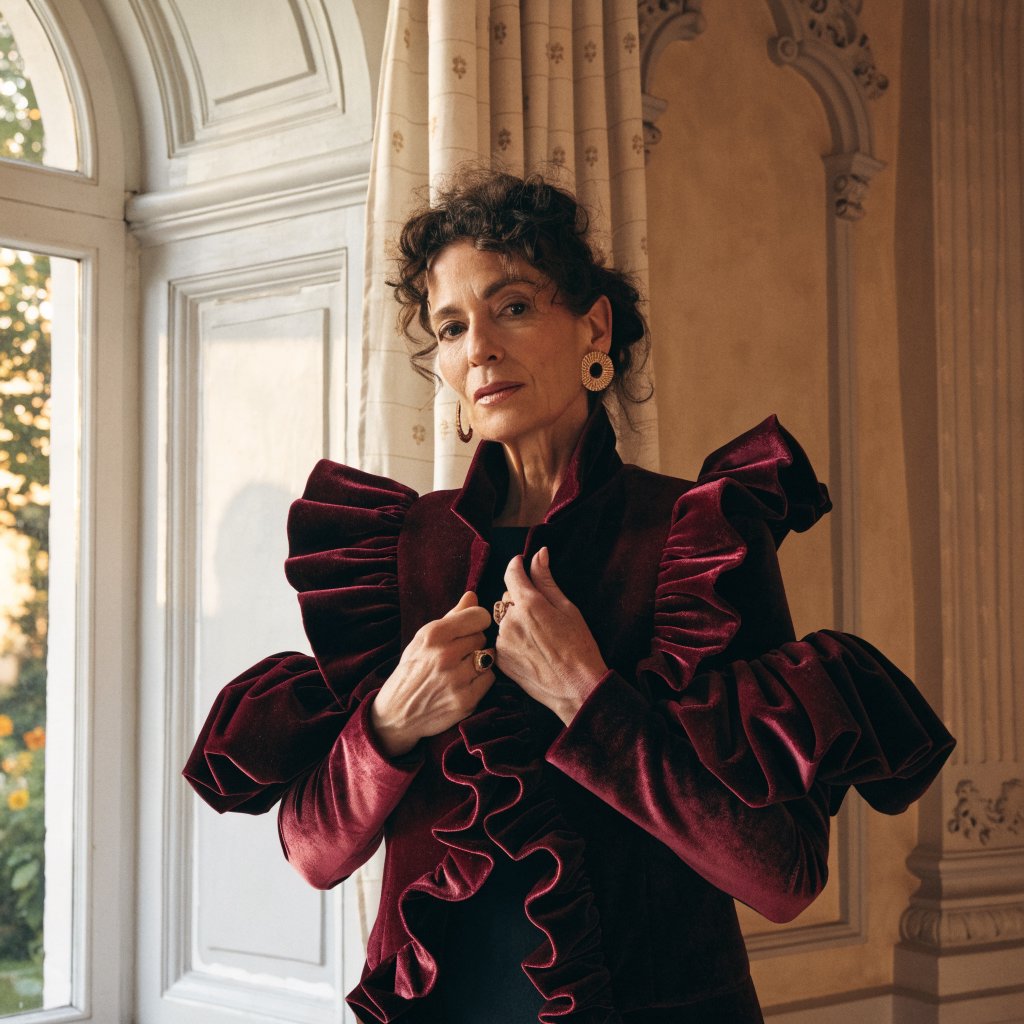 Fashion portrait of an adult woman in her early 50s wearing a sculptural burgundy velvet jacket, her hands lightly gathering its lapel. A wry, composed expression meets the camera against a warm ivory plaster backdrop, lit by soft late-afternoon window glow. — Recraft v4