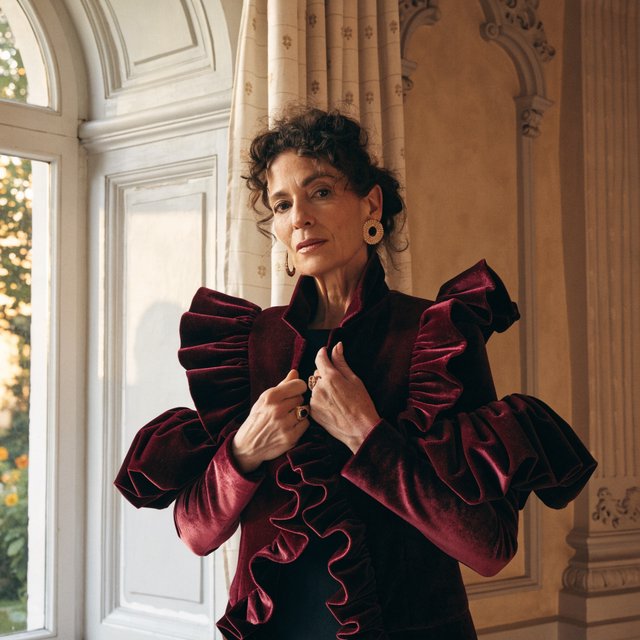 Fashion portrait of an adult woman in her early 50s wearing a sculptural burgundy velvet jacket, her hands lightly gathering its lapel. A wry, composed expression meets the camera against a warm ivory plaster backdrop, lit by soft late-afternoon window glow. — Recraft v4