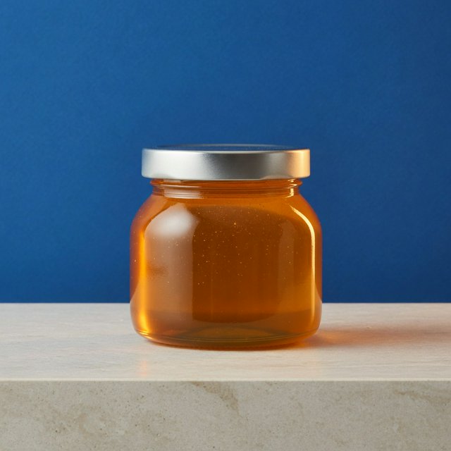 A squat unbranded honey jar with clear glass walls and a plain brushed-aluminum lid rests on honed pale limestone; broad diffused key light and warm rim light shape the commercial product image against a seamless cobalt-blue background, with no lettering. — Model 6
