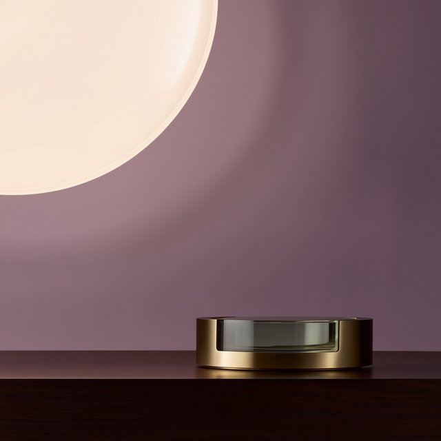 Commercial product photograph of an unbranded sculptural table clock made from satin brass and smoked glass, resting on a dark walnut surface against a muted lavender background, illuminated by a large diffused softbox from the upper left and a subtle rear rim light. — Midjourney Niji 7