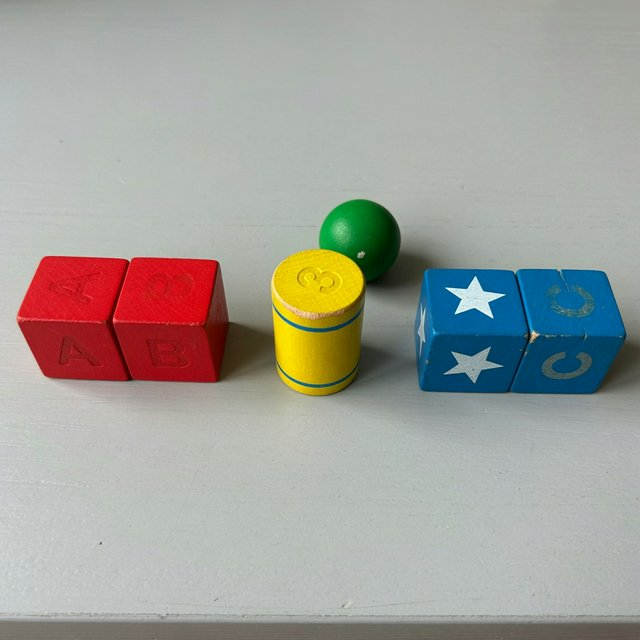 Exactly five wooden toy blocks sit on a pale gray table: two red cubes on the left, one yellow cylinder in the center, and two blue cubes on the right. A small green ball sits behind the cylinder, nothing else. — Grok Imagine Image Quality