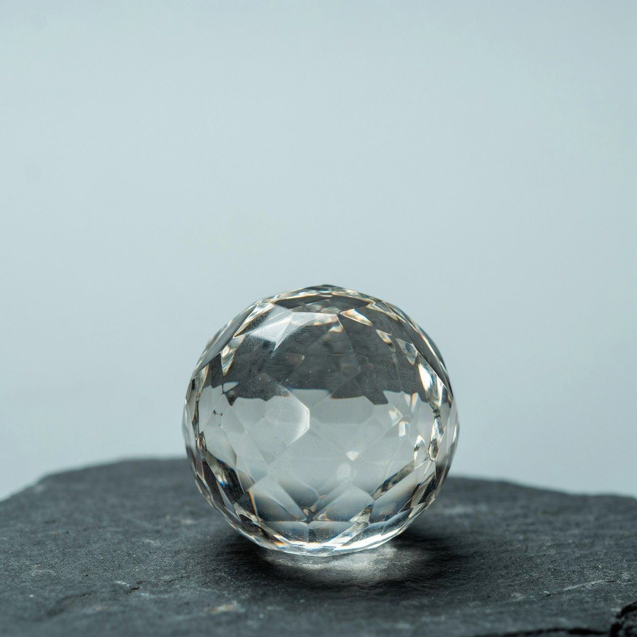 An unbranded, unmarked faceted optical-glass paperweight rests on charcoal slate against a pale blue-gray background; broad diffused studio light and a narrow side highlight reveal crisp internal refractions and bright edge reflections. — Ideogram v4