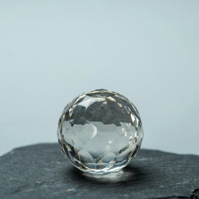 An unbranded, unmarked faceted optical-glass paperweight rests on charcoal slate against a pale blue-gray background; broad diffused studio light and a narrow side highlight reveal crisp internal refractions and bright edge reflections. — Ideogram v4