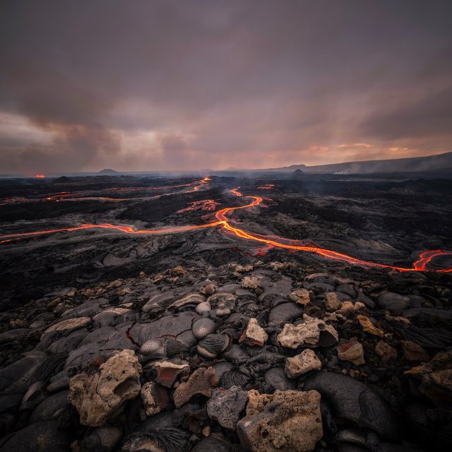 Fresh lava threads a black cinder plain on a volcanic island at dusk beneath ash-laden wind; blistered crust and brittle pumice in the foreground give way to glowing fissures fading into smoke. — Ideogram v4.5