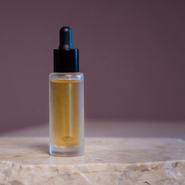 Under a broad diffused key and cool edge light, an unbranded frosted-glass serum bottle displays clear golden oil against a muted plum background, resting on honed pale travertine with its blank surface free of lettering. — Ideogram v4.5