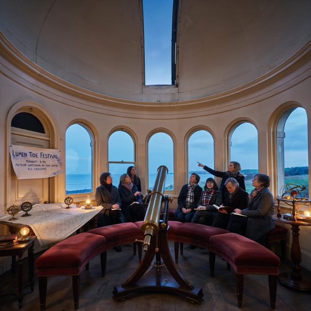 At a restored seaside observatory, a twilight meteor-watching event fills the domed hall with brass instruments, star charts, and velvet benches; "LUMEN TIDE FESTIVAL" is hand-printed in deep blue ink across a cream paper announcement beside the entrance. — Ideogram v4