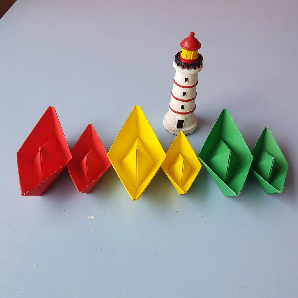 Exactly six folded paper boats sit on a pale blue tabletop: two red boats on the left, two yellow boats in the center, and two green boats on the right. A small white lighthouse figurine stands behind the center pair, nothing else. — Grok Imagine Image Quality