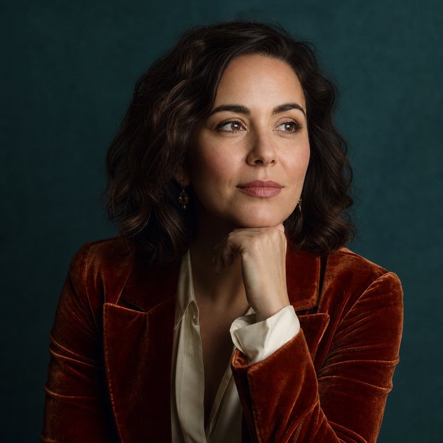 Portrait of an adult woman in her 30s wearing a rust-colored velvet jacket over an ivory blouse, one hand resting beneath her chin, thoughtful expression, deep teal studio backdrop, soft side lighting and gentle cheek highlights. — GPT Image 2