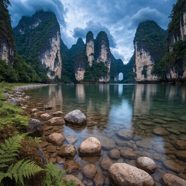 A limestone karst valley at blue hour after a light shower, towering pale cliffs rising above a clear river, low clouds snagging on pinnacles, slick ferns and rounded pebbles leading toward a distant stone arch. — Recraft v4 Pro