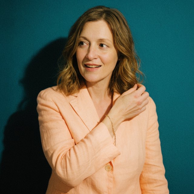 Portrait photograph of an adult woman in her early 30s wearing a pale apricot linen blazer, her hands gently smoothing its cuff. She looks quietly delighted against a deep teal paper backdrop, illuminated by soft, angled studio light. — Krea 2 Large