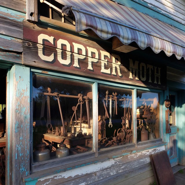 An old roadside bicycle workshop beneath a faded striped awning, rusted tools displayed in the window, with "COPPER MOTH" painted in cream block letters across the weathered wooden fascia, afternoon sunlight catching peeling turquoise paint. — ImagineArt 1.5 Pro