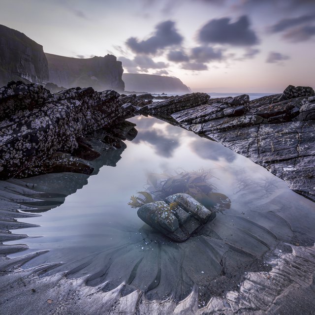 At dawn on a rugged basalt shore, a tide pool mirrors clearing rain clouds; translucent water reveals rippled sand, barnacle-crusted rock, and blurred kelp below the surface, with slick foreground stone leading toward hazy coastal cliffs. — Kling v3