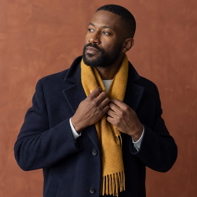 Editorial portrait of an adult Black man in his 30s wearing a deep navy wool overcoat and mustard scarf, fingers adjusting the scarf, thoughtful expression, muted terracotta studio backdrop, crisp cool window light. — GPT Image 2.5 Sunburst