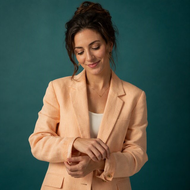 Portrait photograph of an adult woman in her early 30s wearing a pale apricot linen blazer, her hands gently smoothing its cuff. She looks quietly delighted against a deep teal paper backdrop, illuminated by soft, angled studio light. — GPT Image 2