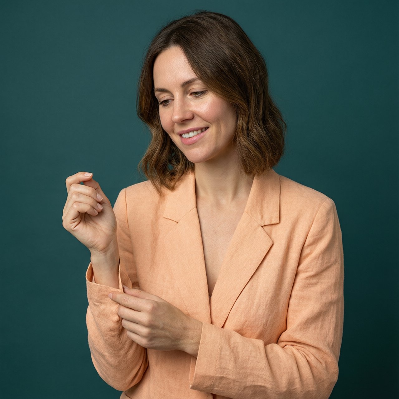 Portrait photograph of an adult woman in her early 30s wearing a pale apricot linen blazer, her hands gently smoothing its cuff. She looks quietly delighted against a deep teal paper backdrop, illuminated by soft, angled studio light. — Qwen Image 3.0 Pro