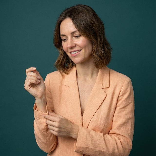 Portrait photograph of an adult woman in her early 30s wearing a pale apricot linen blazer, her hands gently smoothing its cuff. She looks quietly delighted against a deep teal paper backdrop, illuminated by soft, angled studio light. — Qwen Image 3.0 Pro