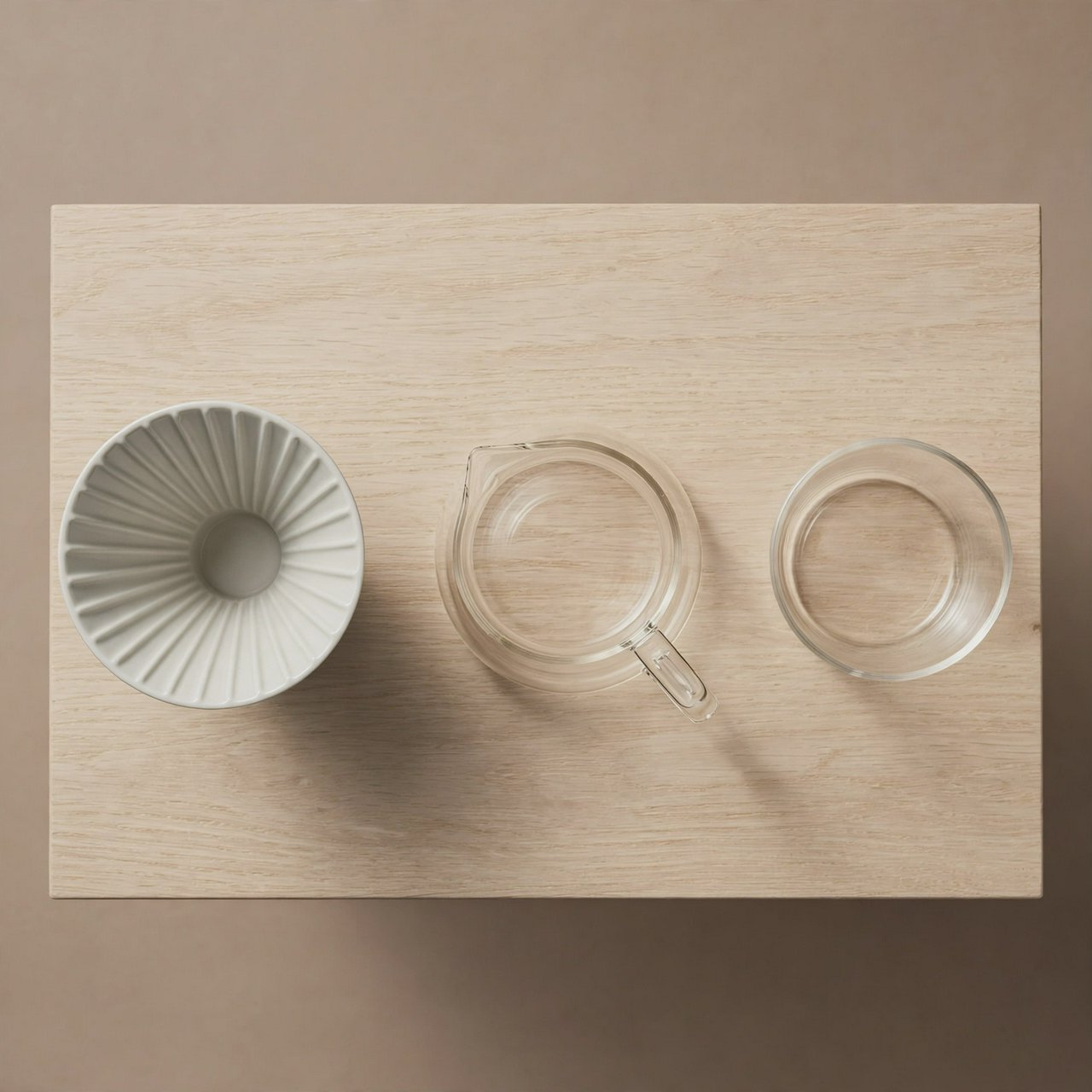 From directly overhead, an unbranded pour-over coffee set with ribbed porcelain and clear borosilicate components arranged in sequence on pale oak; warm taupe background, broad diffused softbox light and gentle side fill shape this commercial product image, no lettering. — Muse Image 1.0