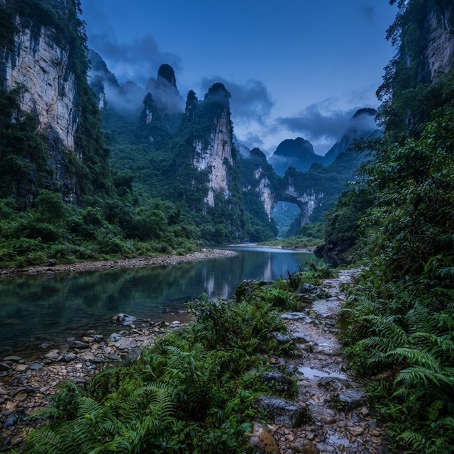 A limestone karst valley at blue hour after a light shower, towering pale cliffs rising above a clear river, low clouds snagging on pinnacles, slick ferns and rounded pebbles leading toward a distant stone arch. — Nano Banana 2 Lite