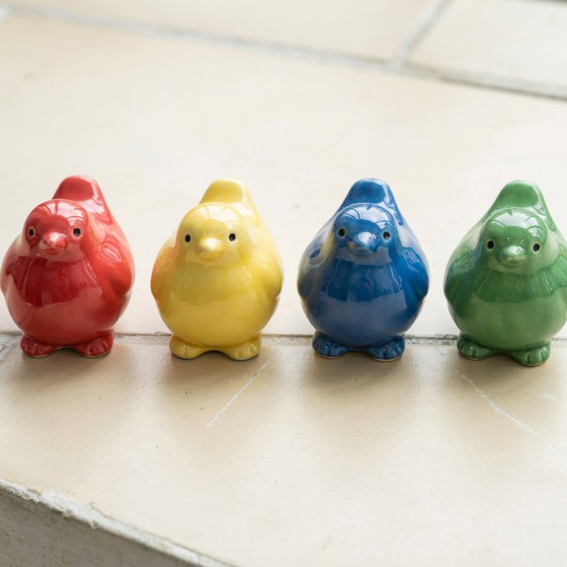 On a sunlit tiled ledge, exactly four ceramic bird figurines form a row: red at far left, yellow at left-center, blue at right-center, and green at far right; all face forward, nothing else. — Ideogram v4.5