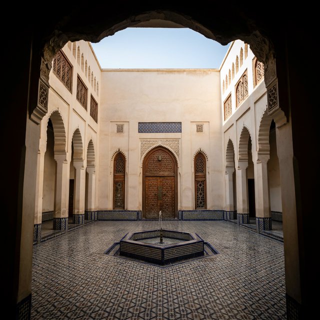 From a shaded courtyard entrance, Moorish Revival residences enclose a tiled fountain court, with cream stucco, carved cedar doors, and blue ceramic bands; late sunlight filters through arcades, revealing aligned walls and credible supports. — Ideogram v4.5