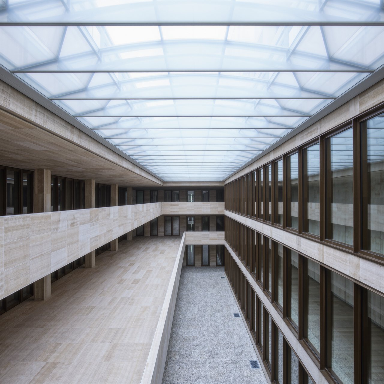 A soaring civic atrium in contemporary Italian Rationalist style pairs pale travertine, bronze mullions, and terrazzo beneath a glazed roof; cool morning light falls from above, viewed from a mezzanine, with aligned bays and credible supports. — Kling v3 Omni